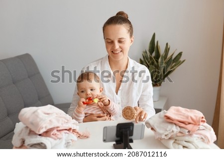Pleasant looking young adult woman posing with infant daughter in front of tripod with phone, filming video for her vlog, talking with her followers about eco toys for kids.