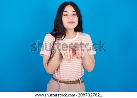 Caucasian woman wearing striped T-shirt over blue background makes bunny paws and looks with innocent expression plays with her little kid