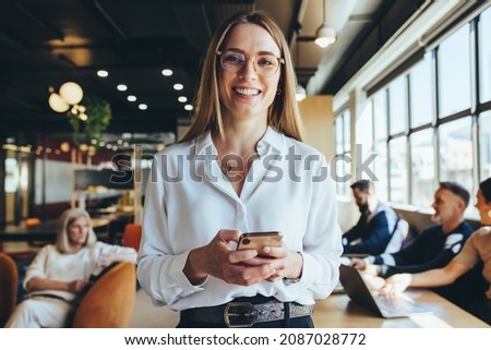 Young businesswoman holding a smartphone in a co-working space. Happy young businesswoman smiling at the camera while standing in a modern workplace. Female entrepreneur sending a text message. Royalty-Free Stock Photo #2087028772