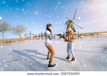 Loving couple having fun on ice in typical dutch landscape with windmill. Woman and man ice skating outdoors in sunny snowy day. Romantic Active date on frozen canal in winter Christmas Eve