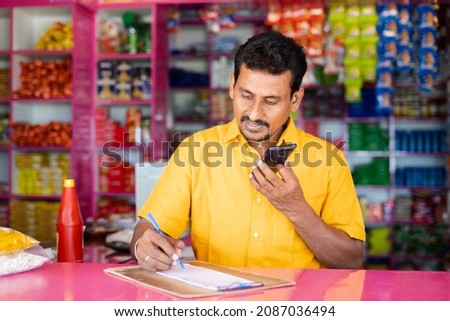 Indian groceries business man noting orders while talking with customer on mobile phone at kirana shop - concept of distance shopping, home delivery service. Royalty-Free Stock Photo #2087036494
