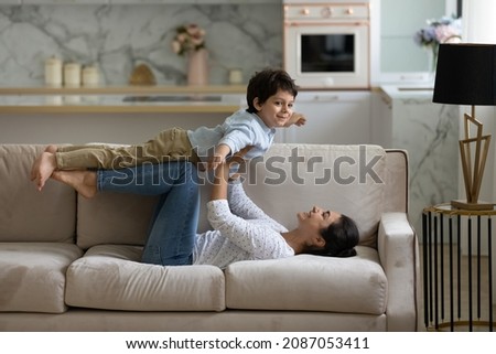 Happy young Indian mother lifting in air on straight arms joyful cute small child son, lying on cozy couch. Joyful family practicing balance doing yoga exercises, having fun playing at home. Royalty-Free Stock Photo #2087053411