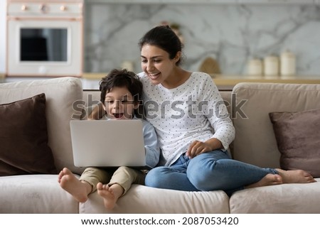 Joyful young Indian woman cuddling laughing small cute kid son, watching together funny comedian movie or cartoons online on laptop, relaxing on couch, tech addiction concept.