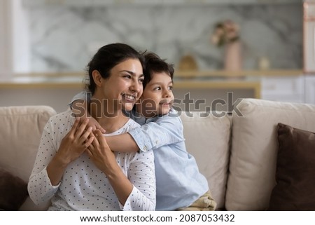 Dreamy bonding sincere small kid boy cuddling Indian young mother, looking in distance, visualizing future or remembering good moments, resting on cozy sofa together at home. Royalty-Free Stock Photo #2087053432