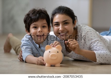 Portrait of happy Indian family lying on floor with small piggybank. Caring young mum teaching little child son saving money for future or planning purchases, financial education concept. Royalty-Free Stock Photo #2087053435