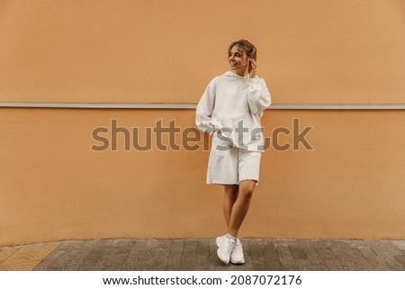 Full-length young Hispanic woman looks to side with legs crossed against orange background. Girl in white sweatshirt with hood, shorts and sports shoes. Elegant style, spring fashion trends.