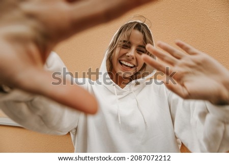 Cheerful young fair-skinned lady hiding from paparazzi hiding behind her hands against background of wall. Blonde with perfect smile wears white sweatshirt. Freedom concept Royalty-Free Stock Photo #2087072212