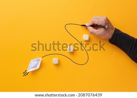 Male hand draws a curved line that guides a paper boat to cross the barriers. Guidance, assistance, leadership or to overcome obstacles concept. Royalty-Free Stock Photo #2087076439