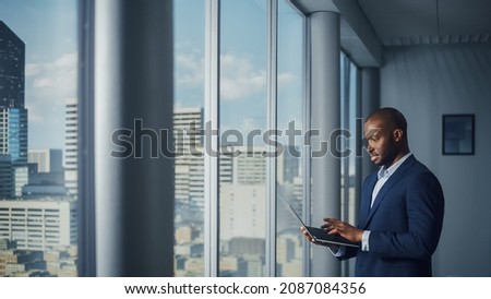 Thoughtful Black Businessman in a Tailored Suit Using Laptop while Standing in Office Near Window on Big City. Successful Corporate Top Manager Doing Data Analysis for e-Commerce Startup Royalty-Free Stock Photo #2087084356
