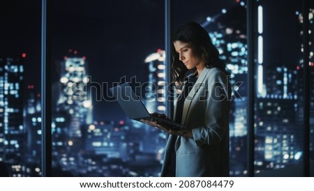 Big City Modern Office at Night: Successful Young Businesswoman Standing and Using Laptop. Beautiful Female Digital Entrepreneur Thinking of Investment Strategy for e-Commerce Project. Royalty-Free Stock Photo #2087084479