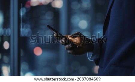 Night Office: Successful Black Businessman Wearing Suit Standing, Using Smartphone. CEO Browsing Internet, Managing Social Media Strategy in e-Commerce Software. Focus on Hand with Mobile Phone Royalty-Free Stock Photo #2087084509