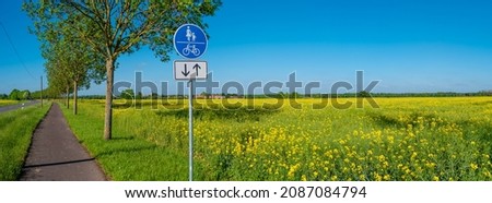 Panoramic view over beautiful farm landscape with rapeseed blossom field and a cycling and hiking lane or path in Germany, Spring, at sunny day and blue sky, with a signpost for bicycles and people