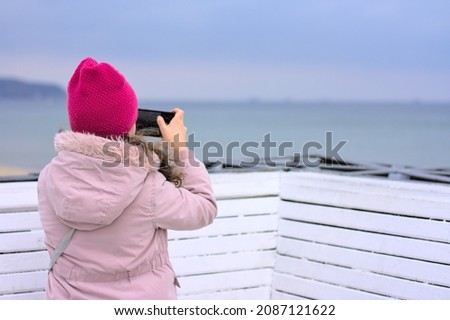 Girl taking a picture of the sea with her phone. Autumn, cloudy day, pier.