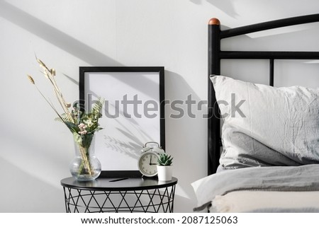 Picture frame with blank space on side table near bed in cozy bright bedroom. Nightstand with alarm clock, mockup art, home decor and flower in vase in room with morning light