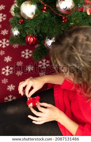 A beautiful young woman in a red sweater holds a box with a gift tied with a red ribbon at home near a Christmas tree on a warm blanket. Selective focus. Close-up