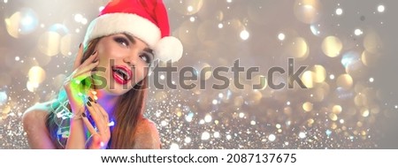 Christmas woman. Beauty model girl in Santa Claus hat with red lips and xmas garlands in her hand looking up. Joy. Surprised expression. Closeup portrait over winter holiday blinking snow background 