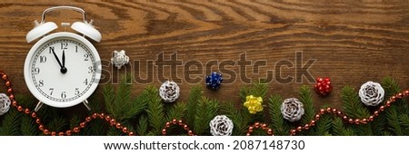 White alarm clock, cones, fir twigs and beads garland on brown wooden table background. Wide banner. Empty place for text. Countdown to midnight. Counting last moments before Christmas or New Year.