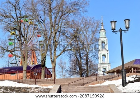 The picture shows a city park with an attraction called a Ferris wheel, a church bell tower and a street lamp.