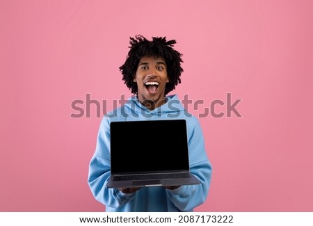 Excited black teen guy holding laptop computer with empty black screen on pink studio background, mockup for website. Cool African American youth demonstrating PC with space for online advertisement