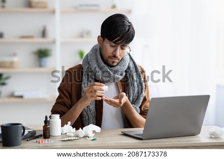 Sick young hindu guy with scarf around his neck taking medicine while sitting in front of laptop at home, looking for treatment against cornavirus, cold or flu on Internet, copy space Royalty-Free Stock Photo #2087173378