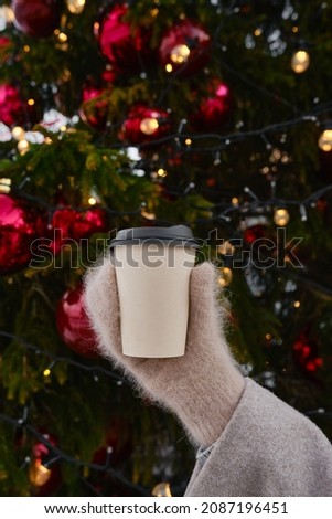 Craft paper cup with coffee, cocoa or tea in the hands of a girl against the background of a Christmas tree. Time to drink hot holiday drinks. Empty space for text, layout. Coffee to go