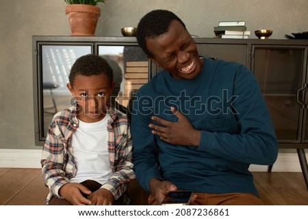 Astonished little preschool boy with brown skin looking at camera with jaw dropping after hearing adult joke from his father sitting next to him on floor laughing out loud, holding phone in hands Royalty-Free Stock Photo #2087236861