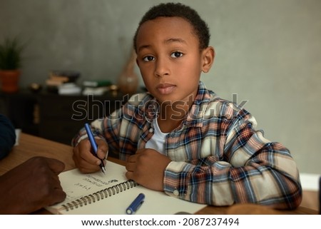 Portrait of adorable dark-skinned honor student doing homework, studying math, writing down numbers, doing sums, looking at camera with smart face expression. Clever child. Childhood and school