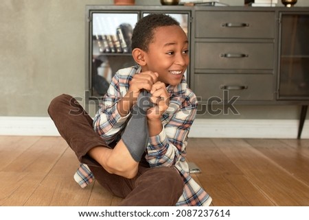 Funny picture of African American male kid taking off his dirty socks after returning home from school, sitting on floor, telling story with enthusiasm, isolated on living-room interior background