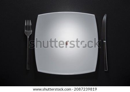 Photograph of a white plate with a bean in the center and metal cutlery on the sides on a black background. Concept photo about poverty, financial problems, lack of food, diet, etc.