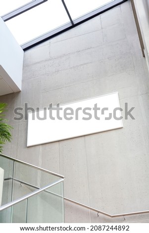 Empty blank white rectangle advertisement frame hanging on a concrete wall in a modern corporate office corridor, nobody. Business building interior staircase wide picture frame template, mock up.