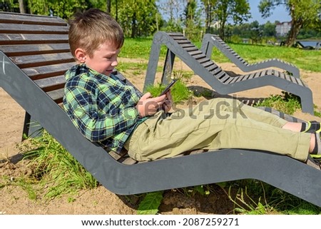 Happy little boy is sitting on a bench and playing on a mobile phone against the background of a summer park.