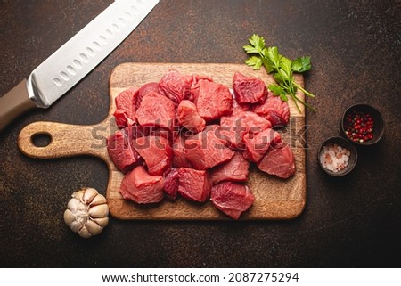 Raw beef meat chopped in cubes with bunch of fresh parsley, garlic, salt and pepper on wooden cutting board with knife for cooking stew or meat dish on brown dark stone background top view flat lay Royalty-Free Stock Photo #2087275294