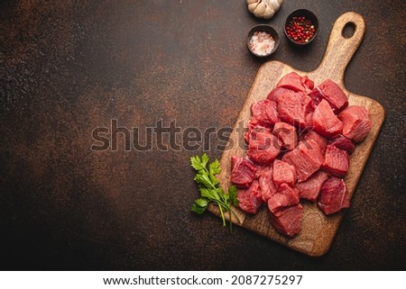 Raw beef meat chopped in cubes with bunch of fresh parsley on wooden cutting board for cooking stew or other meat dish on brown dark stone concrete background top view flat lay space for text Royalty-Free Stock Photo #2087275297