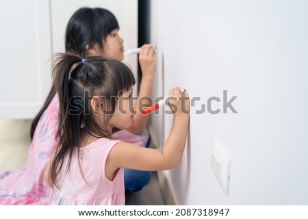 Asian young sibling kid girl enjoy paint on white wall in living room. Little adorable children having fun drawing and coloring art picture with hapiness enjoy creativity activity on holiday at home.