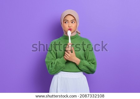 Hungry beautiful Asian woman in green sweater holding a spoon in mouth think of tasty food isolated over purple background