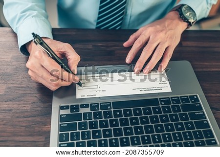 Business man hand writing and signing white blank bank check book and dollar bill, coin, laptop and graph chart on the desk at office. Payment by check, paycheck, payroll concept.
