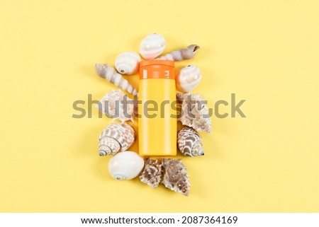 Sun cream with seashells around lies in the middle of a light yellow background with copy space on the sides, top view close-up.