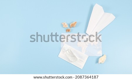 White paper plane, medical mask, disinfectant, world map, and seashells lie on the right against a blue background with a copy of the space on the left, top view close-up.