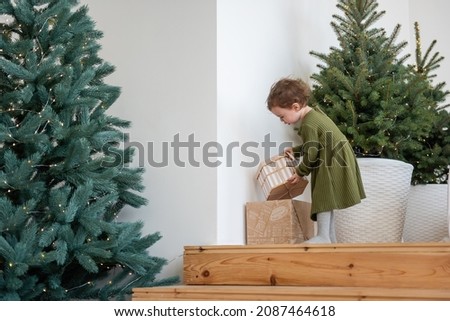 Little girl in warm green dress stands on the wooden steps by the Christmas tree, opens boxes with gifts in the loft living room. Minimalist scandinavian style of the apartment. Festive lifestyle