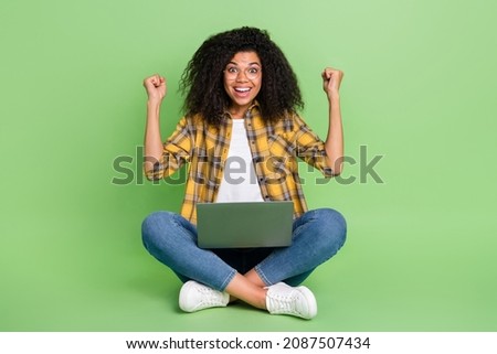 Full length photo of cool curly hairstyle young lady sit hold laptop wear spectacles yellow shirt jeans footwear isolated on green background