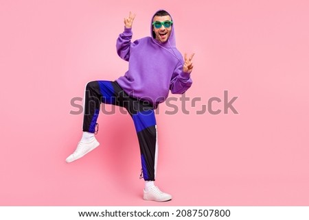 Full length photo of cool young brunet guy dance show v-sign wear eyewear hoodie pants shoes isolated on pink background