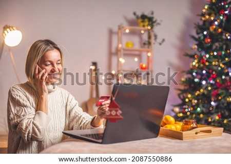 Happy adult woman shopping online on Christmas, using laptop, phone and credit card, xmas sales concept. Smiling middle aged lady talking on cellphone, sitting at home and buying xmas gifts 