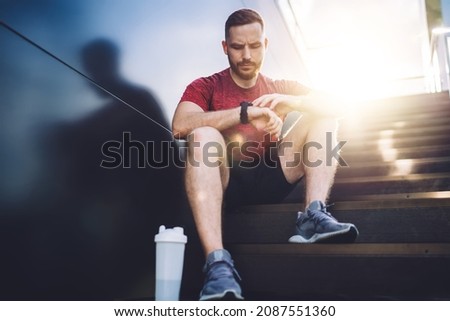 Low angle of thoughtful man in sneakers looking at smartwatch after doing jog exercise on street in sunny summer day