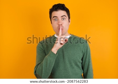 Surprised caucasian man wearing knitted green sweater isolated over yellow background  makes silence gesture, keeps finger over lips and looks mysterious at camera