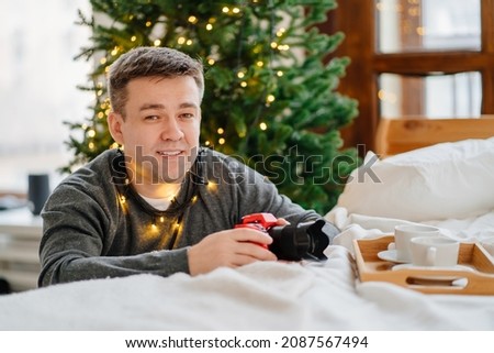 man in gray sweater with garland with camera in red silicone case by bed against background of Christmas decorations. work of photographer during Christmas holidays. accessories for equipment.