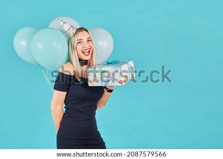 Beautiful smiling woman in black dress and birthday hat holding gift box and pastel air balloons against blue background. Cute happy young girl celebrating birthday party.
