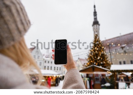 A girl holds a smartphone in her hands and takes pictures of a decorated Christmas tree on the street to share photos on social networks. Festive mood and atmospheres. Christmas tree