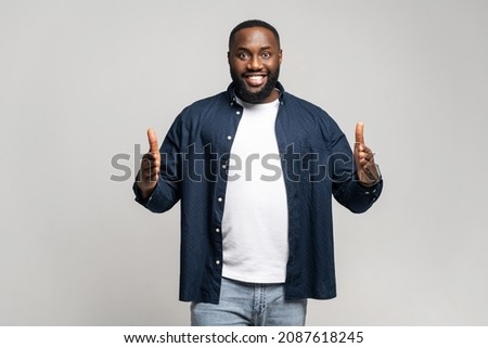 Positive African man with beard looks at camera, holding two hands in front of him showing the size on gray isolated background in studio, wearing casual shirt, smiling joyfully, presenting something