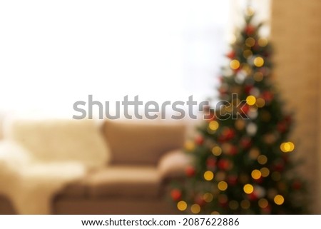 Blurred out traditional christmas pine tree with holiday decorations. Decorative lights glowing on fir with bokeh effect. Close up, interior background with beige couch, copy space for text.