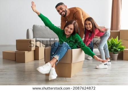 Celebrating Moving Day. Portrait of happy Arab family having fun together in new apartment, unpacking, smiling husband riding excited wife in cardboard box in living room with little daughter Royalty-Free Stock Photo #2087624872